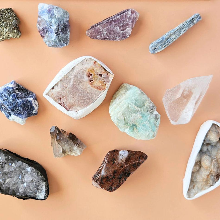 A calming crystal set displayed on a neutral wooden tray, featuring five polished stones—lepidolite, howlite, amethyst, amazonite, and smoky quartz—partially spilling from a cotton pouch beside a handwritten card labeled “Peaceful Mind Crystal Kit.”